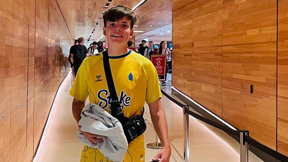 Mackenzie has short brown hair and is wearing a yellow Everton shirt and a cross body bag. He is stood inside Sydney airport.