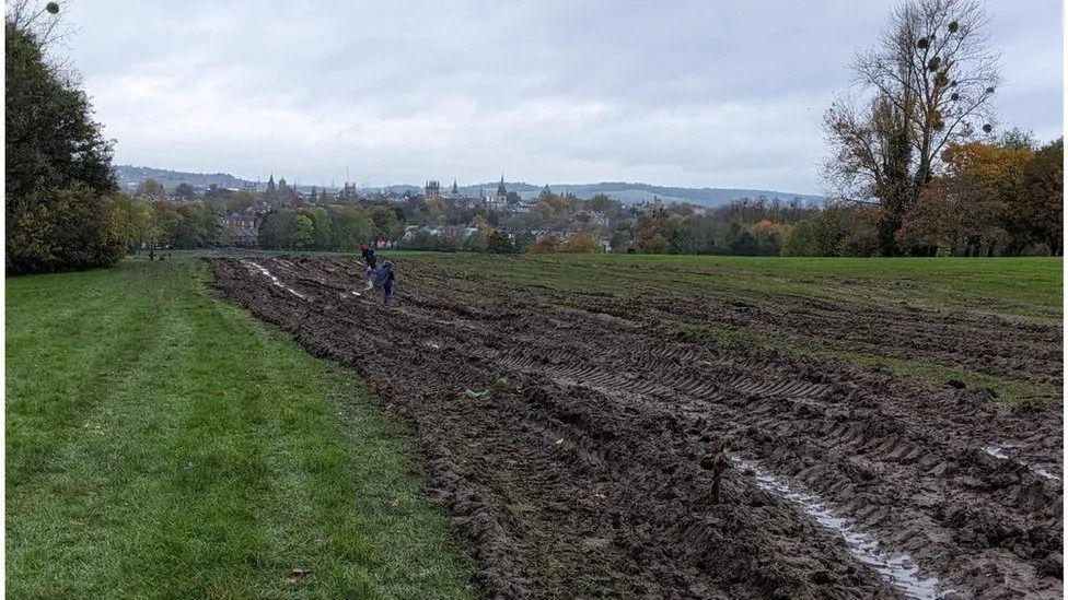 A muddy field with tyre tracks buried into the ground and people working to fix the damage