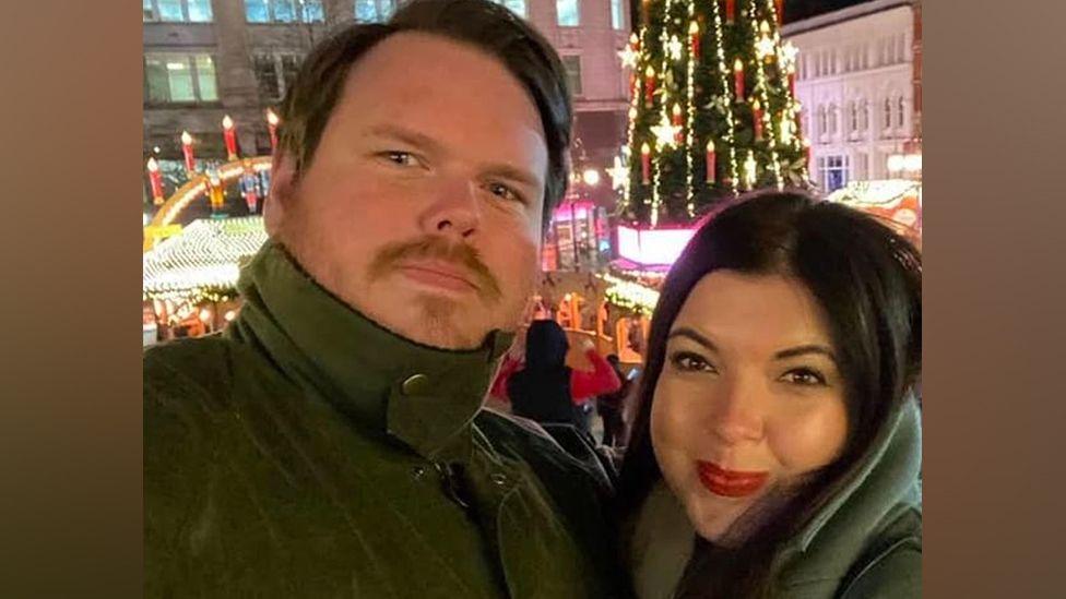 Steven (left) and Hannah Haggerty are taking a selfie. There is a lit up Christmas tree in the background. Steven Haggerty has brown hair and a brown moustache. He wears a green anorak buttoned up to his chin. Hannah Haggerty has black hair and is smiling with a closed mouth. She is wearing a green coat.