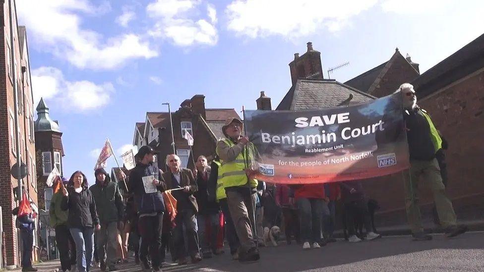 A group of protestors walking through a street with banner saying "Save Benjamin Court"