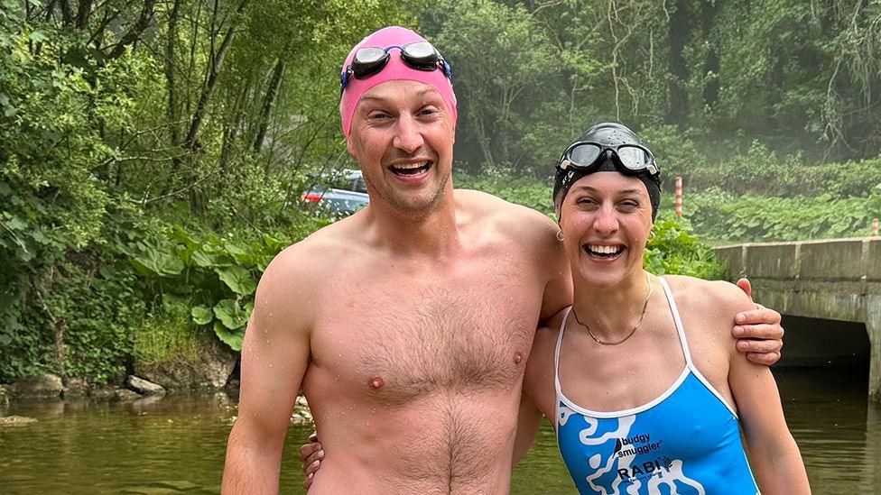 Brother and sister Hugh and Alex Addison are standing in a river with a concrete bridge behind them, wearing swimming costumes and smiling broadly. He has a pink swimming cap and she has a black one. Both have swimming goggles resting on the top of their heads.