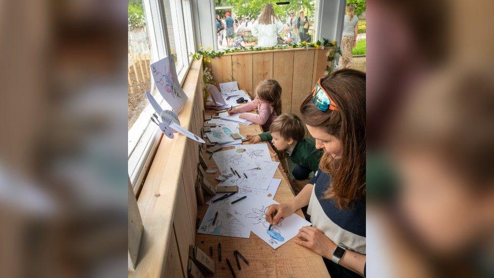 A young boy, a young girl, and an adult woman sitting at a wooden bench colouring in pictures of bees