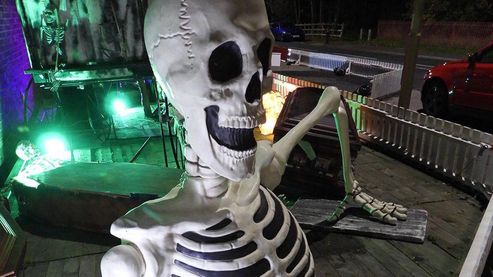 A close-up image of a plastic skeleton, perched on a bench, with more skeletons arranged in the background in a Halloween display, featuring a coffin and coloured lights.