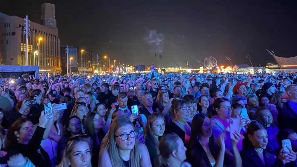 A huge crowd, many holding mobile phones, on the prom. In the background you can see the town lit up in the distance and a big wheel