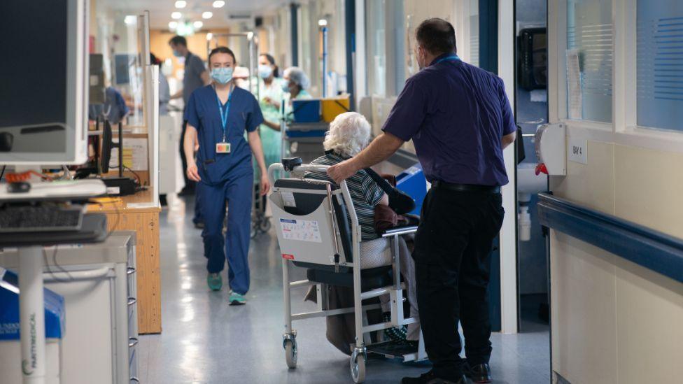 Patients and staff pictured in a hospital corridor, with an elderly women in a wheelchair in the foreground 