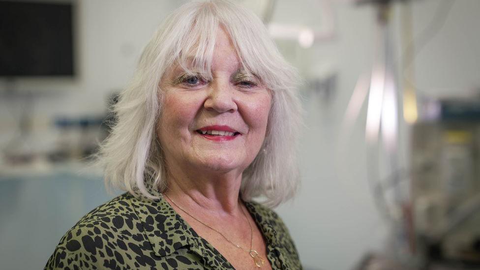 Doreen Adams, woman with shoulder-length grey hair and middle parting, smiling, wearing a green and black blouse and a necklace, with medical equipment in the background.