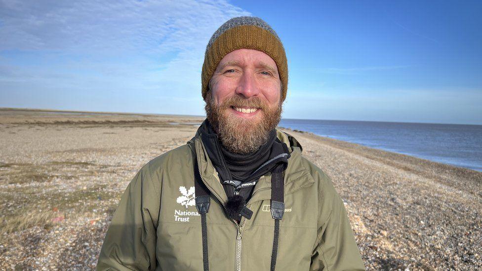 Matt Wilson smiles at the camera while standing on a shingle beach. He is wearing a green coloured beanie hat and a green coat. He has a light coloured beard.