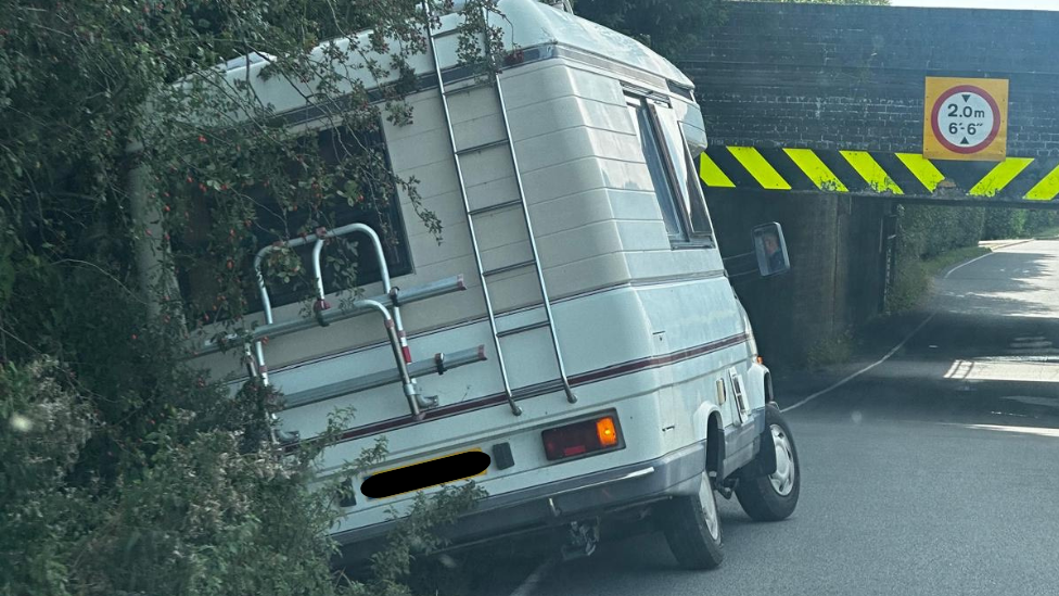 A white and beige camper van in a verge at the approach to the 2m (6ft 6inch) high underpass.
A low 2m (6 ft 6 inch) high underpass, with circular red and white warning signs that has unseen railway track above.
The is a yellow and black chevron that is damaged by vehicles hitting.