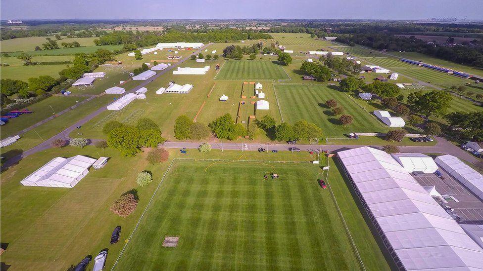 An aerial view of Trinity Park in Ipswich. The showground is set up for the Suffolk Show, but people are yet to be let in. We can see several arenas with patterned, mown grass as well as temporary white tents or varying sizes, roadways, and some trees. The rest of the countryside is visible on the horizon, as are the cranes at the Port of Felixstowe.