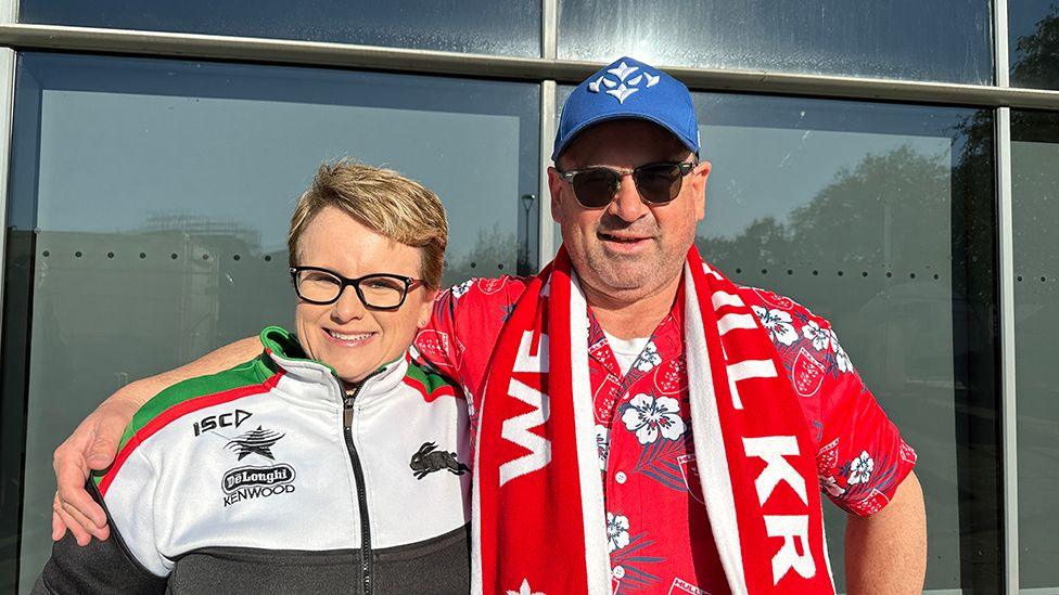 Leonard Gregory dressed in sunglasses, a red Hawaiian shirt, blue baseball cap and Hull KR scarf stands with his arm around his wife who is wearing a black and white tracksuit and dark glasses 