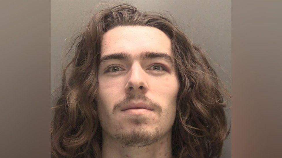 A mugshot of Connor Chapman who has long curly hair, a brown moustache and wispy stubble, and smirks at the camera 