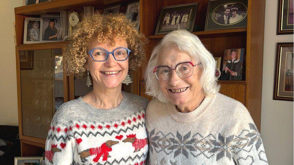 Clare is on the left with her mum on the right. Clare is wearing stripy blue classes and has ginger curly hair. She is wearing a knitted jumper with dogs on it. Her mum is wearing glasses and has white hair and is wearing a white and grey knitted jumper. They are both smiling.