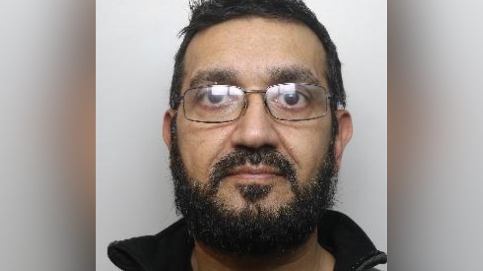 A police mugshot of Mohammed Khan. He has short dark hair and a beard. He is wearing glasses.