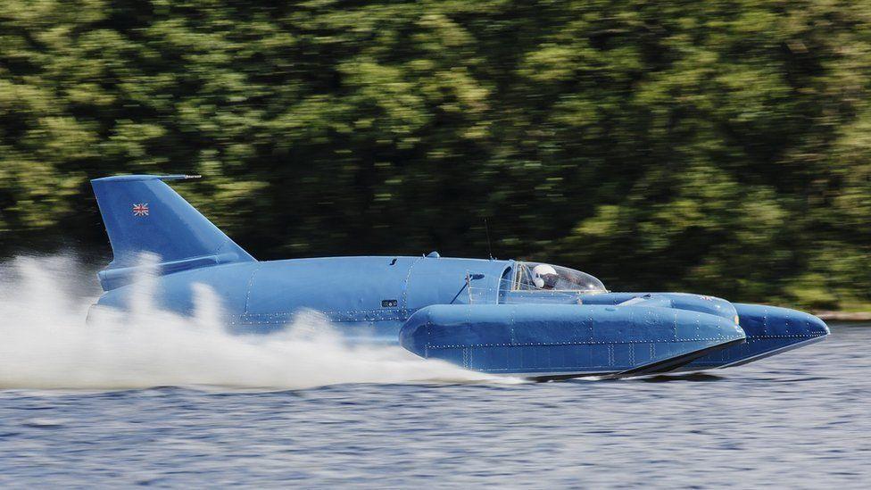 Bluebird, a jet-engined craft painted blue, running on a lake. Spray from the water is shooting up behind it. The pilot's white crash helmet and dark visor are visible in the cockpit.