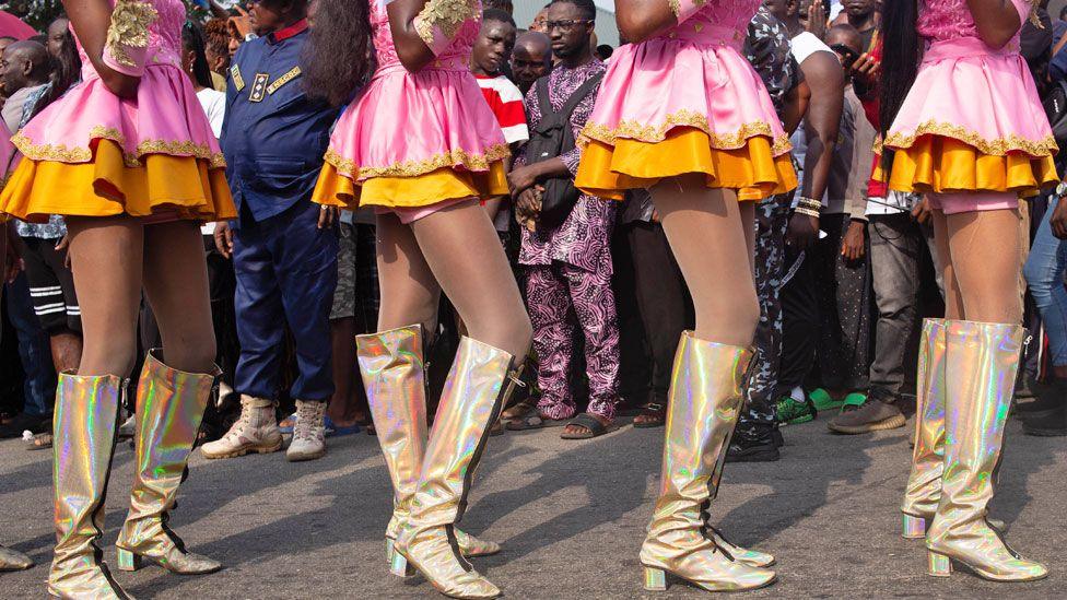 Parade onlookers look through the legs of performers in gold boots and pink and orange