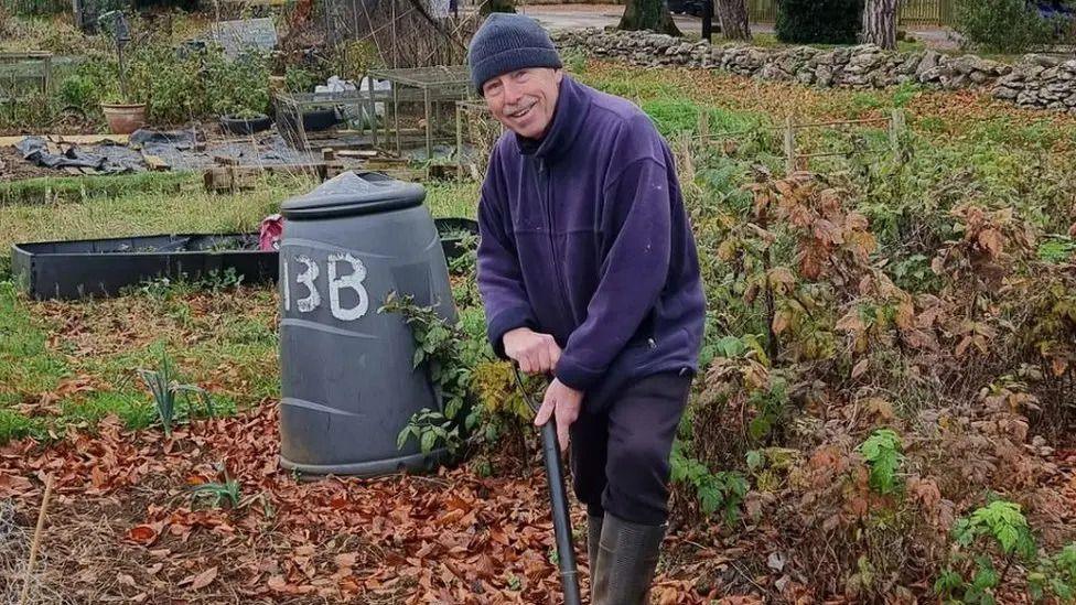 Chris Pearce is standing in his allotment. He is raking leaves. He is wearing a blue fleece and a hat.