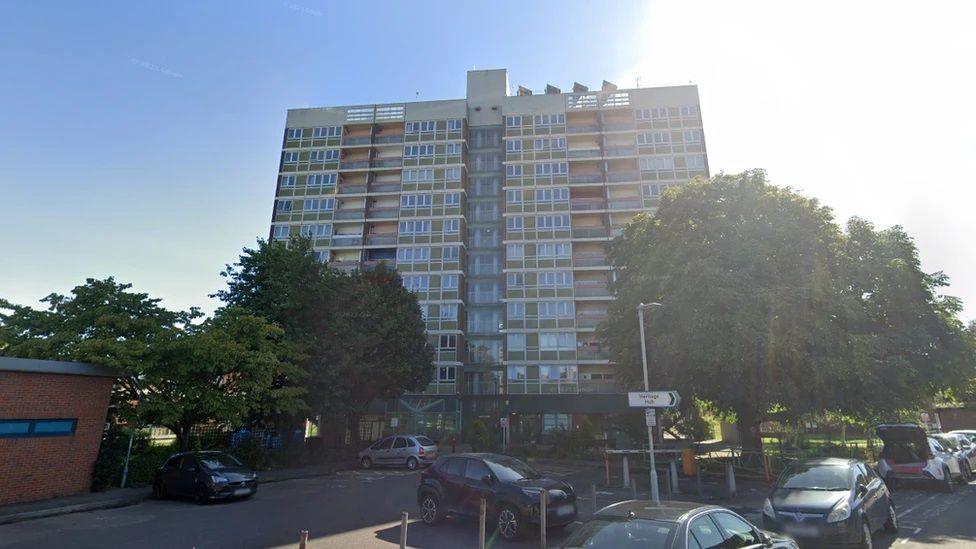 A high-rise tower block called Clapham Court in Gloucester with some trees and parked cars in front of it. The picture is taken on a sunny day with blue sky