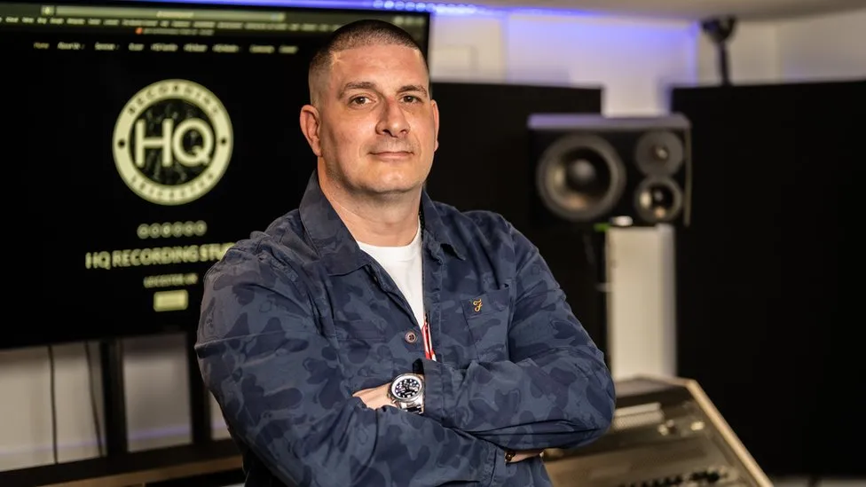 A man with a blue jacket on and his arms crossed looking at the camera. He is standing in front of a screen that says HQ Recording Studios