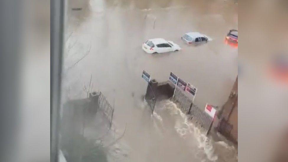 Submerged cars can been under brown flood water, which has smashed through black gates into a car park. 