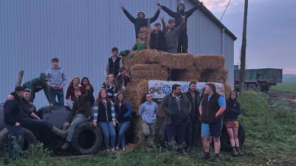 A straw bale sculpture covered in young men and women. Some are standing on it, some are standing in front of it and others are sitting or standing to its side. Behind them is a grey barn and the back end of a trailer.