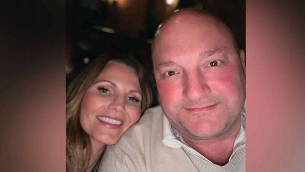 Lucy with her head on Rob's shoulder in a close up selfie. He wears a cream shirt and beige v-necked jumper and they are both smiling.