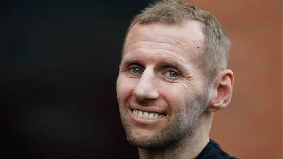 Rob Burrow smiling at the camera with short brown hair, a brown beard and black top. The background is black and brown too.