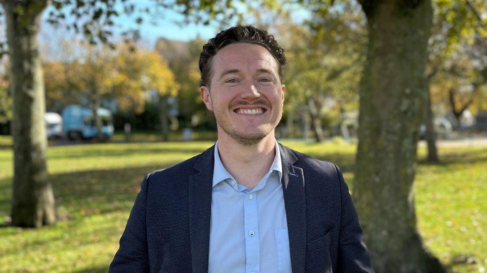 A man stood in a green park. He is smiling and wearing a light blue shirt and a dark navy blazer. There are some trees behind him as well as lots of grass.