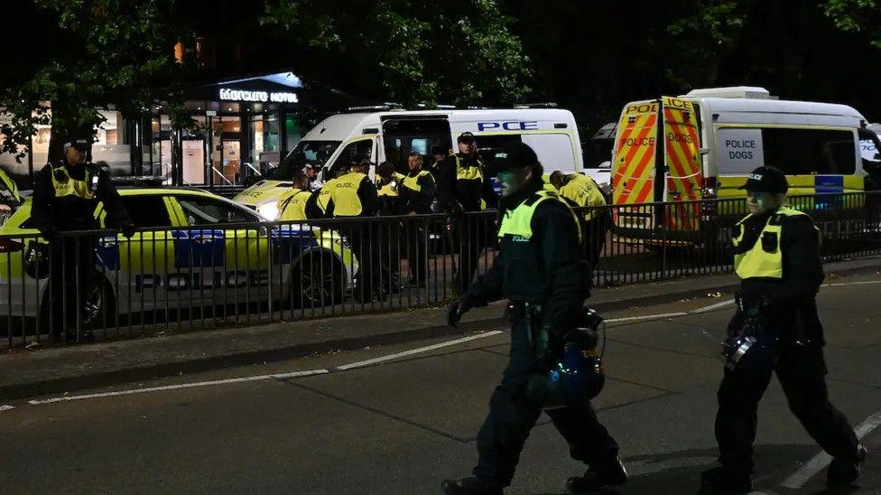 Two police officers walking on a road in the foreground with more police cars and vans on the other side of the road.