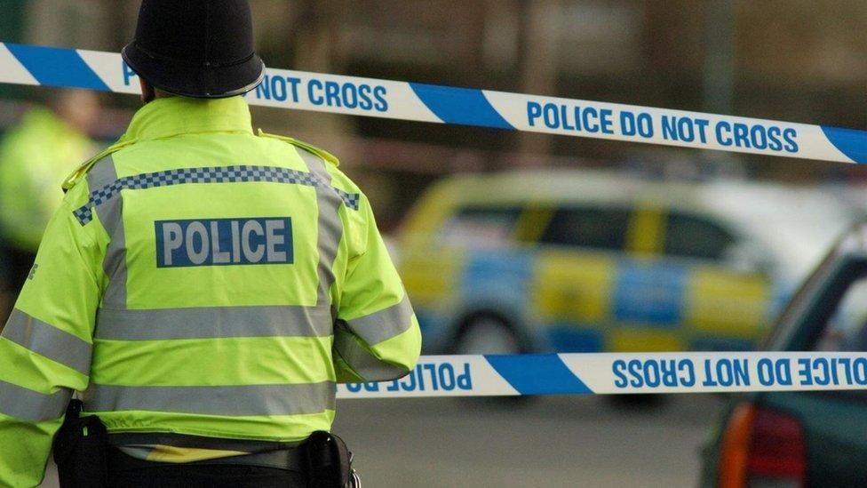 A police man on the left wearing a yellow hi-vis jacket and a police hat. He is approaching two lines of blue 'do not cross' police tape which is cordoning off an area. In the distance you can see a blurred out police car parked on the street.