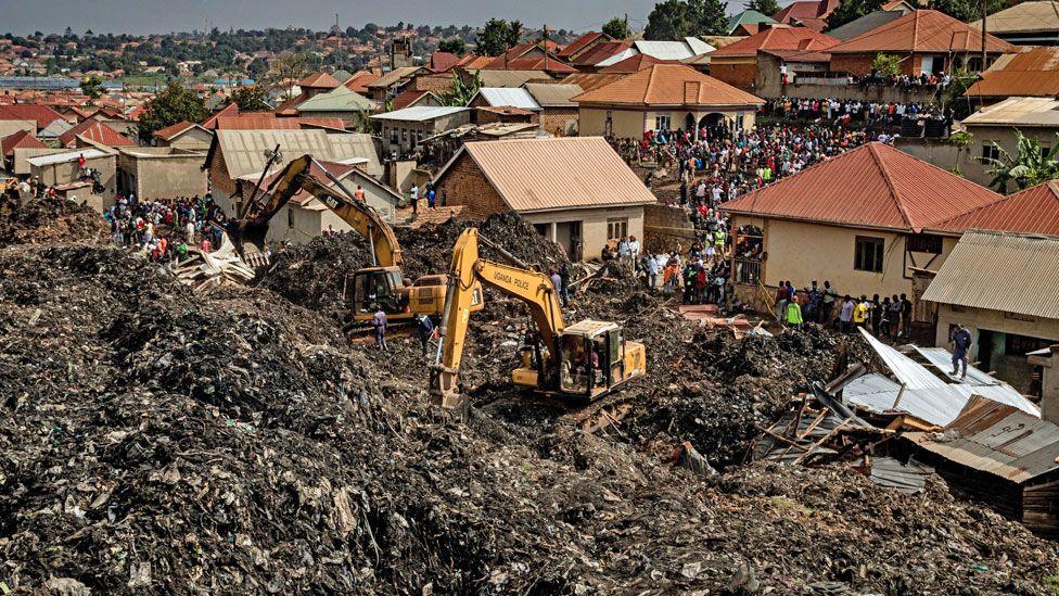 Two yellow diggers are seen amidst the rubbish of Kiteezi with houses seen behind them and a crowd of onlookers - Kampala, Uganda, August 2024.