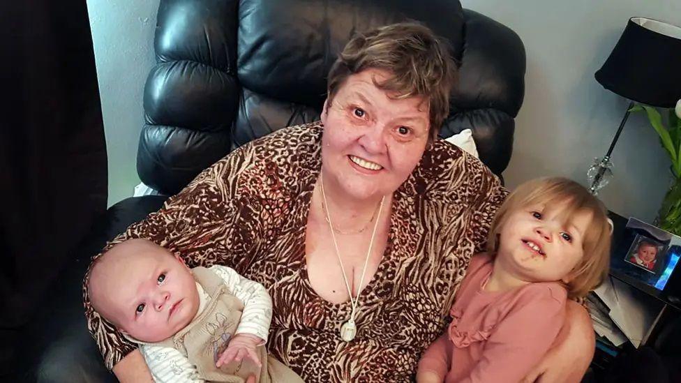 Wendy is wearing an animal-print top with a long gold necklace. She is sat in an armchair cuddling one baby and another child and is smiling up at the camera.