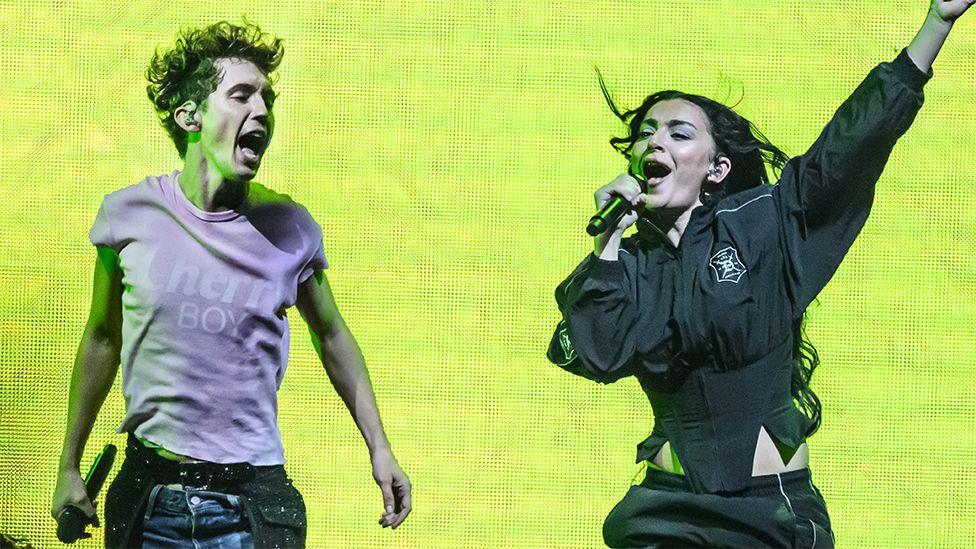 Troye Sivan and Charli XCX on stage performing. Troye, a man, is on the left wearing a pink tshirt jumping up, while Charli is in black singing into a microphone with her left arm outstretched. Behind them is a lime green electronic backdrop.