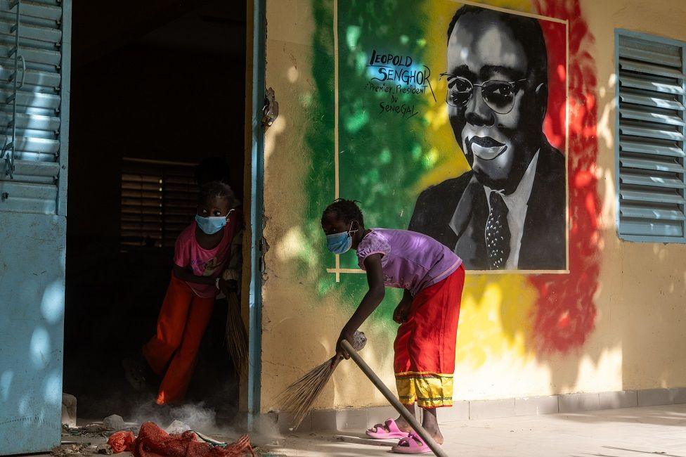 Children sweep the floor at Medina Gounass Elementary School. On the wall is a mural of the country's first president, Léopold Sédar Senghor.