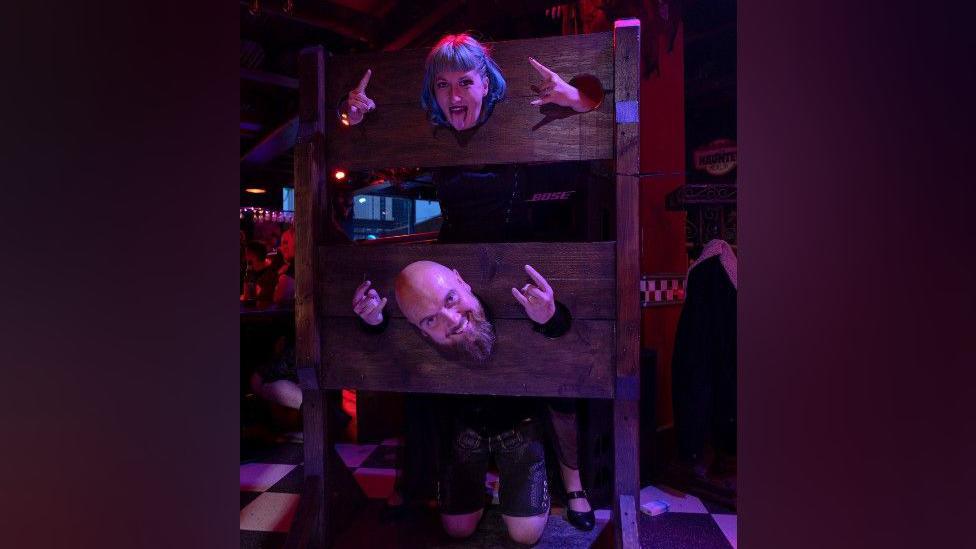 A woman with blue hair is standing in medieval-style wooden stocks, with her head and both hands through circular holes. She has her tongue out and is doing "rock" signs with her hands. Below her is a man who is bald and has a long beard. He is on his knees with his head and both hands through the stocks' holes. He is smiling while holding his hands in "rock" signs