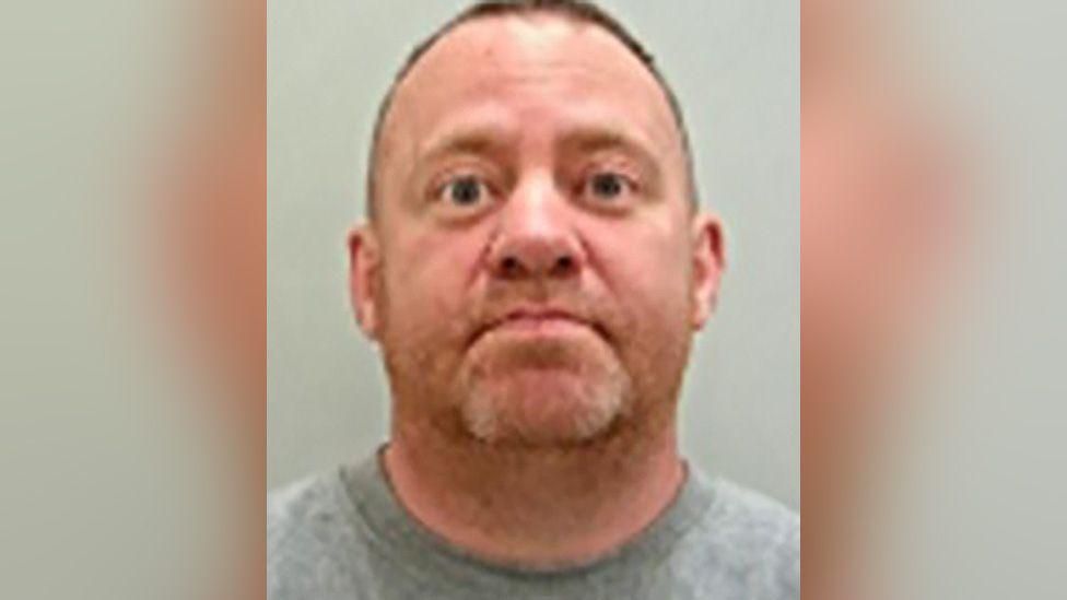 Police custody image of Neil Reid, who has short brown hair and blond stubble. He is wearing a grey t-shirt.