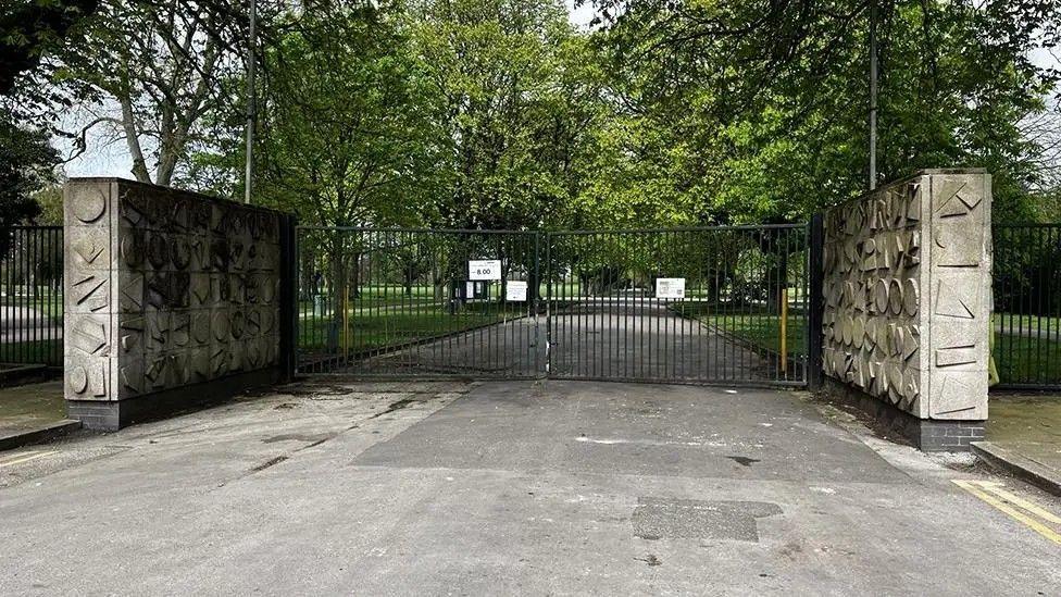 A gated entrance to a park with a long tree-lined avenue in the background. There are two ornate walls to either side of the black gates, which are closed.