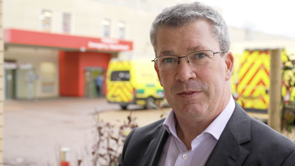 Joe Harrison is clean shaven, wearing a pink shirt and dark-coloured suit jacket. He is wearing glasses. Behind him is a hospital building and ambulances