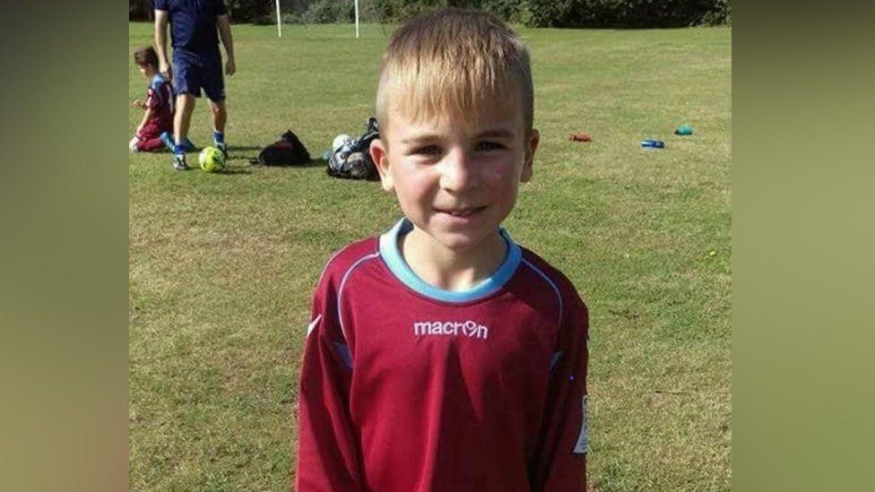 Kieron Hilling as a young boy, wearing a claret and blue football kit. He is smiling at the camera.