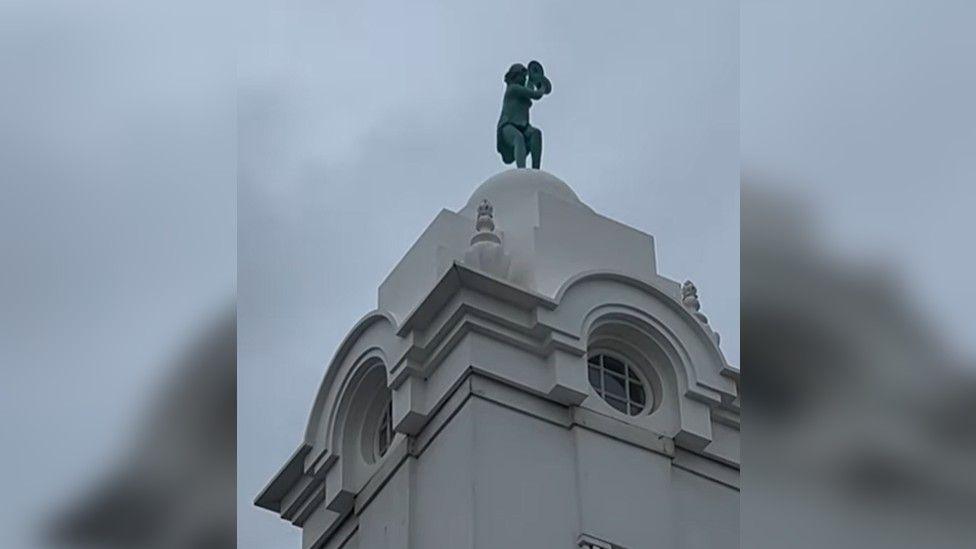 A close up of the small, green dancing lady statue on top of a white domed tower with circular windows. The sky is grey and cloudy.