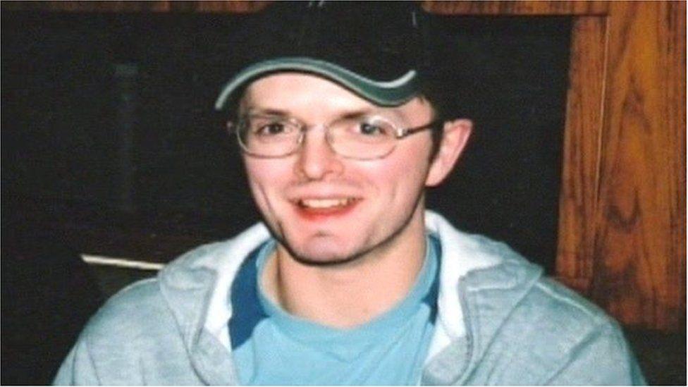 Paul McCauley is a young man. He is smiling at the camera. He is wearing small wire-rimmed glasses and a dark baseball cap. He is wearing a light blue t-shirt under a gray hooded top.  