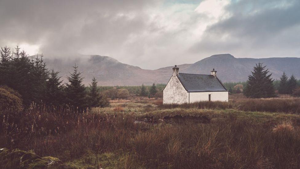 A small white house sits in a rural landscape surrounded by trees and grass with hills in the background and an ominous cloudy sky