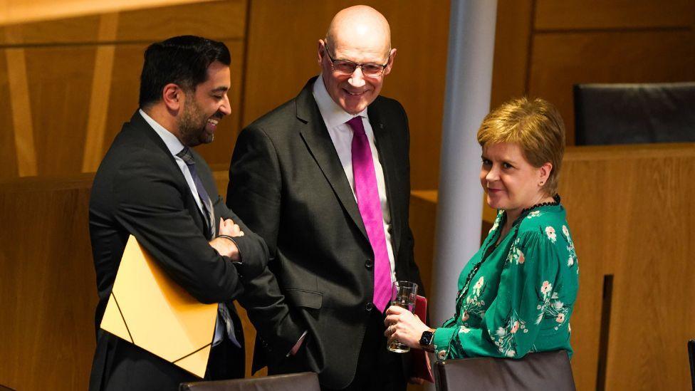 Yousaf, with dark hair, Swinney, who is bald and Sturgeon, with light-coloured hair, stand in the Scottish Parliament chamber. Sturgeon, wearing a green top is leaning on a chair looking away from the men, who are standing next to each other smiling, wearing dark suits. 