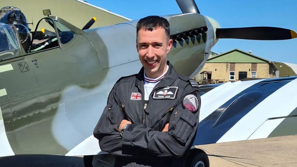 Sqn Ldr Mark Long in dark blue RAF flying suit standing with arms folded in front of a Spitfire aircraft