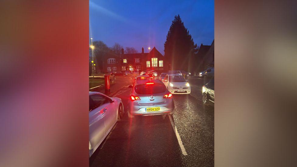 Cars are pictured on a residential road at night, stuck in traffic. The view is behind one car, and two cars facing each other appears to be trying to squeeze past each other with two parked cars on either side of them