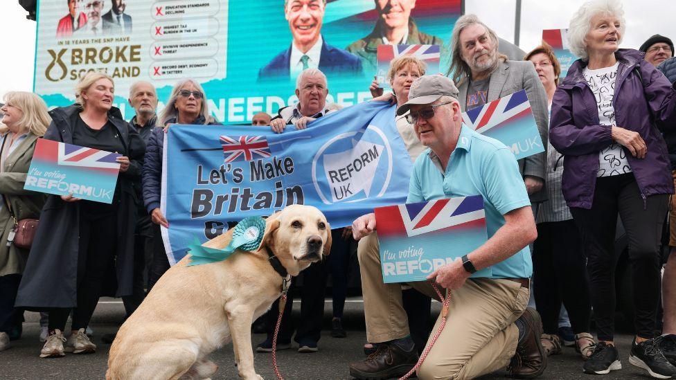 A group of people holding Reform signs and banners line up behind a large flag. There man with a Reform sign and a labrador dog with a Reform rosette in the foreground, and a large Reform billboard in the background.