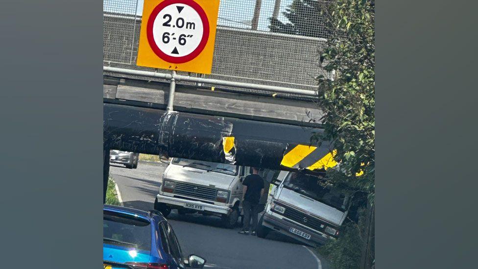 A white van stuck under the Stonea Road Bridge with another one at its side, falling into the verge. A man is standing with his back to the viewer between the vans. The bridge has a sign above it by saying it is 2m or 6ft6in tall