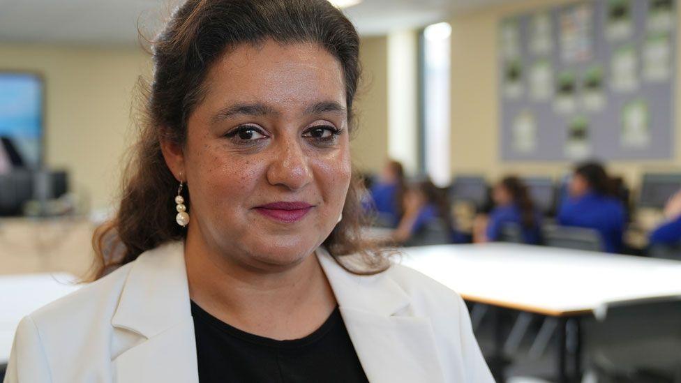 Shahneila Saeed, who has black long hair pulled back from her forehead. She is wearing a cream jacket over a black top and starting to smile. She is standing in a classroom, which is blurred out but tables and pupils in bright blue stops can be seen. 
