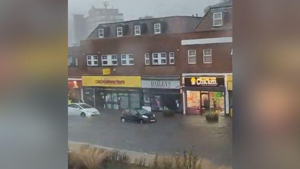 Cars driving on Dunstable High Street, which has been covered in floodwater due to heavy rain. There are shops in the background.