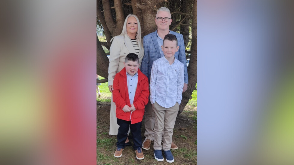 Alfie (bottom left) with his family. A woman behind him has blonde hair, wearing a beige coat and black and white striped top. A boy beside him is wearing a a light blue shirt, beige chinos and blue trainers. Alfie is wearing a red coat, light blue shirt, black trousers and brown shoes. His dad Jamie is wearing black glasses, a blue check blazer and blue shirt. They are all standing in front of a tree.