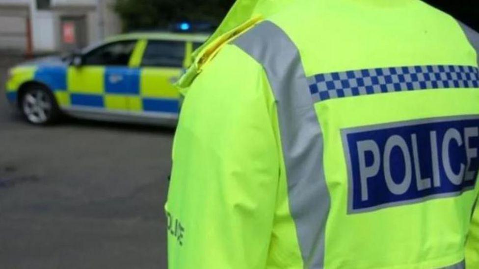 A shot of a police officer taking from behind. The officer wears a hi-vis jacket and stands in front of a police car.